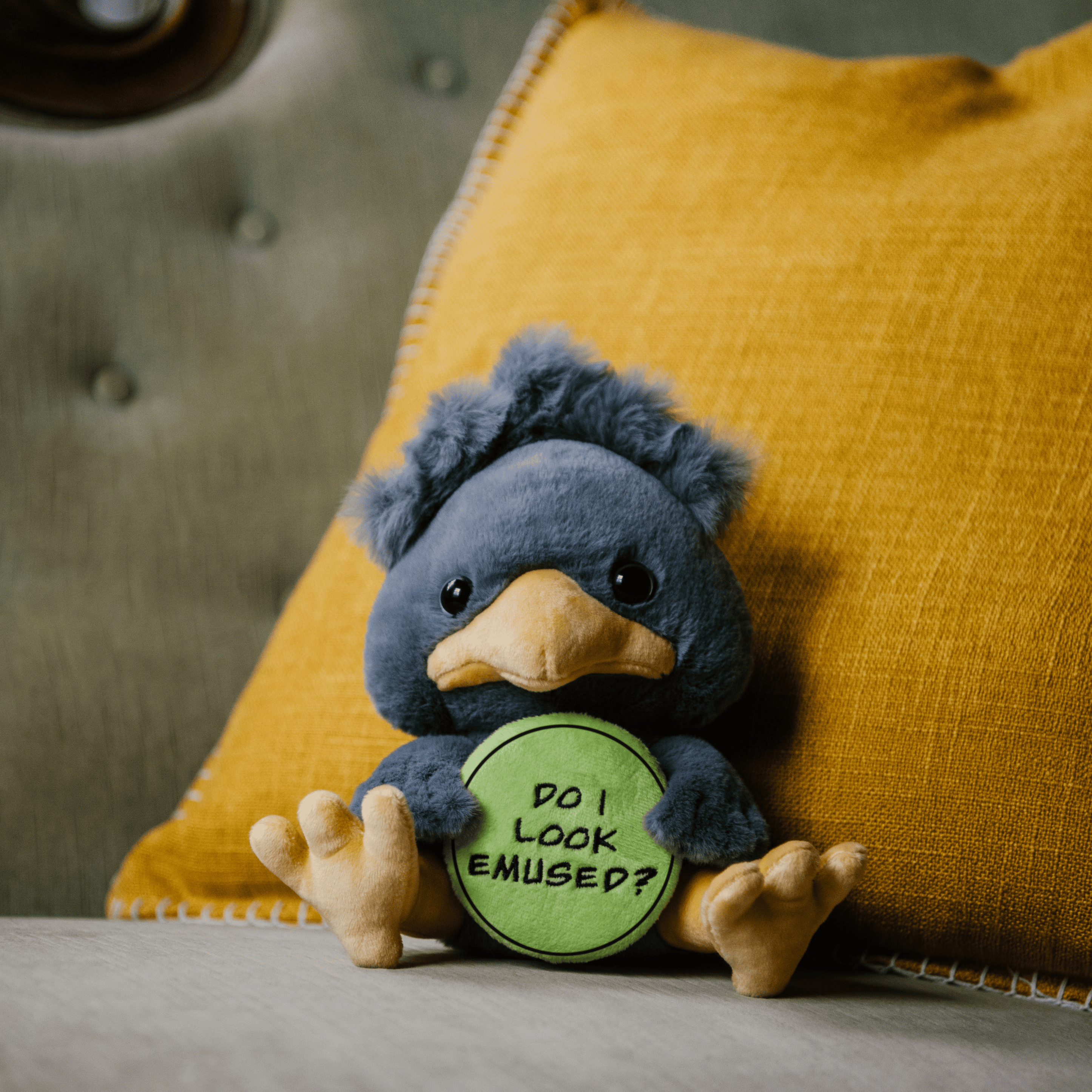 A plush depicting a gray emu holding a green sign with the text 'DO I LOOK EMUSED?' placed on a couch.