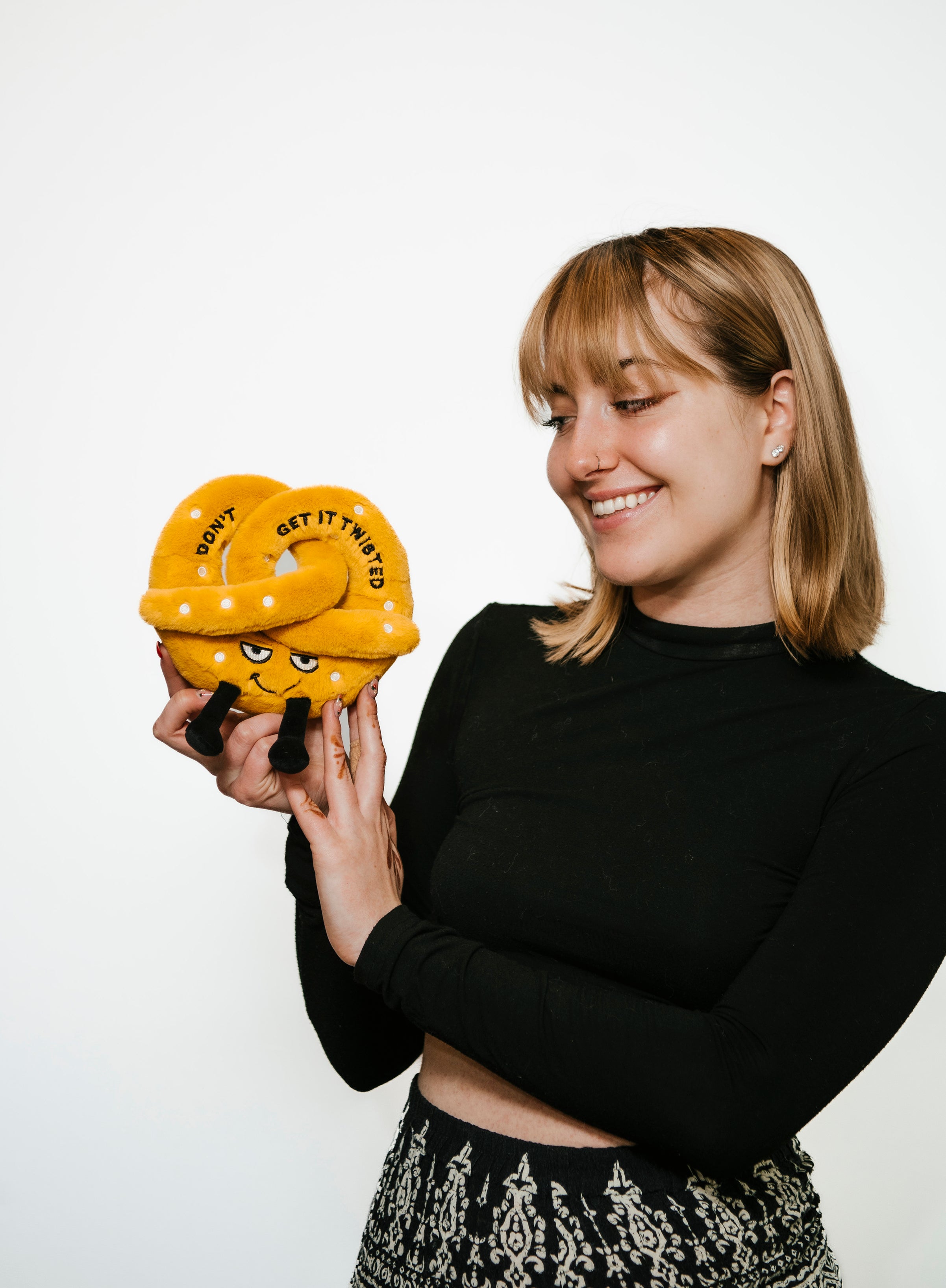 A woman holding a plush designed to look like a twisted pretzel with a grumpy facial expression, dangly legs, and topped with simulated salt. It reads, 'Don't get it twisted.'