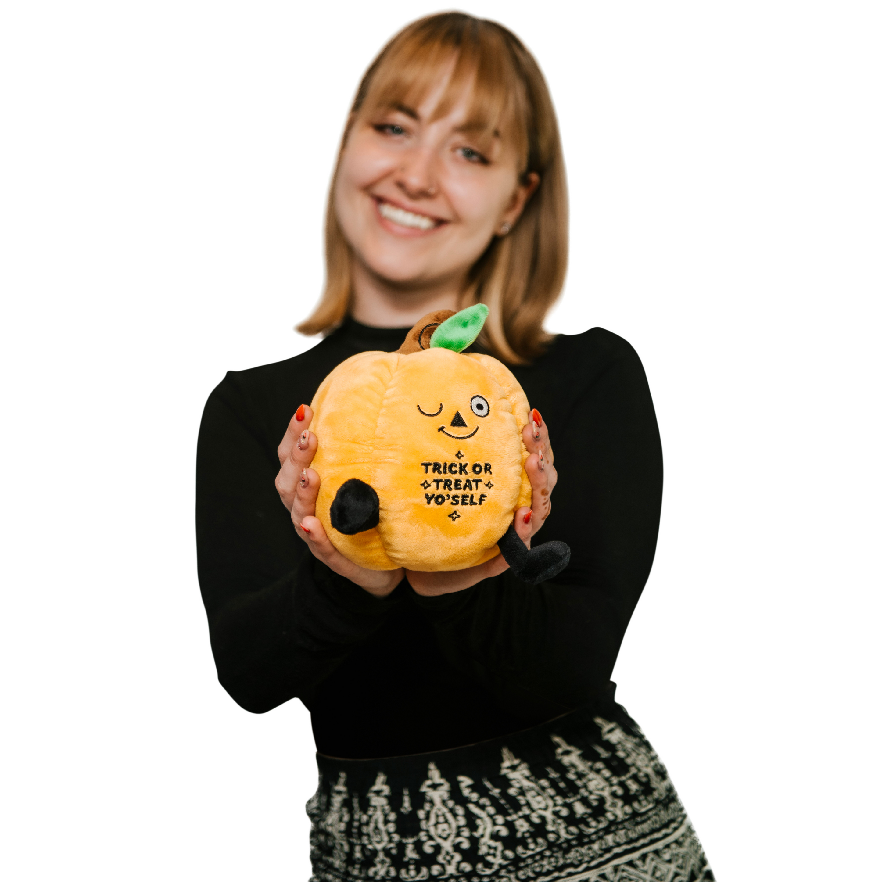 A woman holding a plush designed to look like a smiling orange pumpkin with the phrase 'TRICK OR TREAT YO'SELF' written on it.