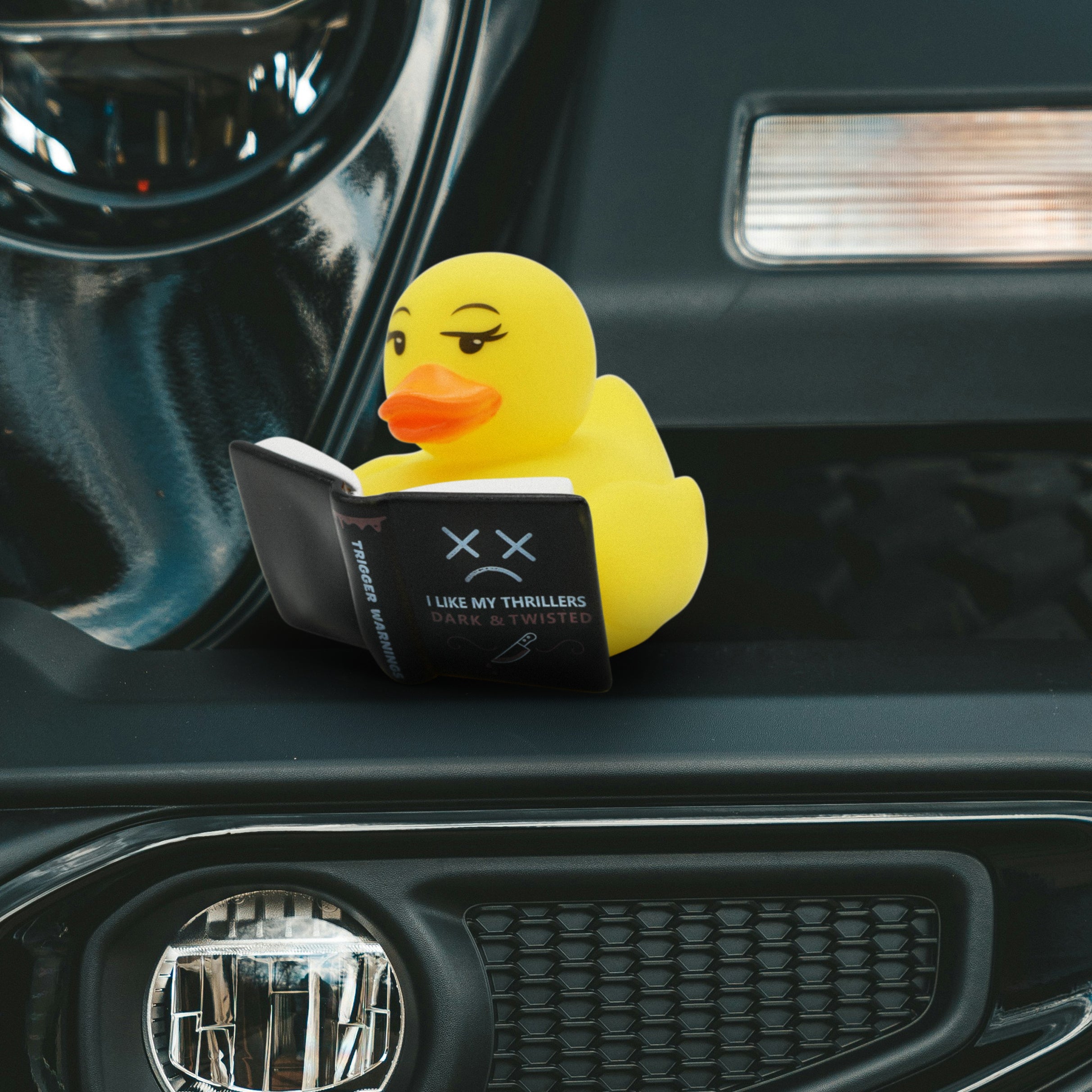 Yellow rubber duck reading a thriller book placed on a car dashboard.