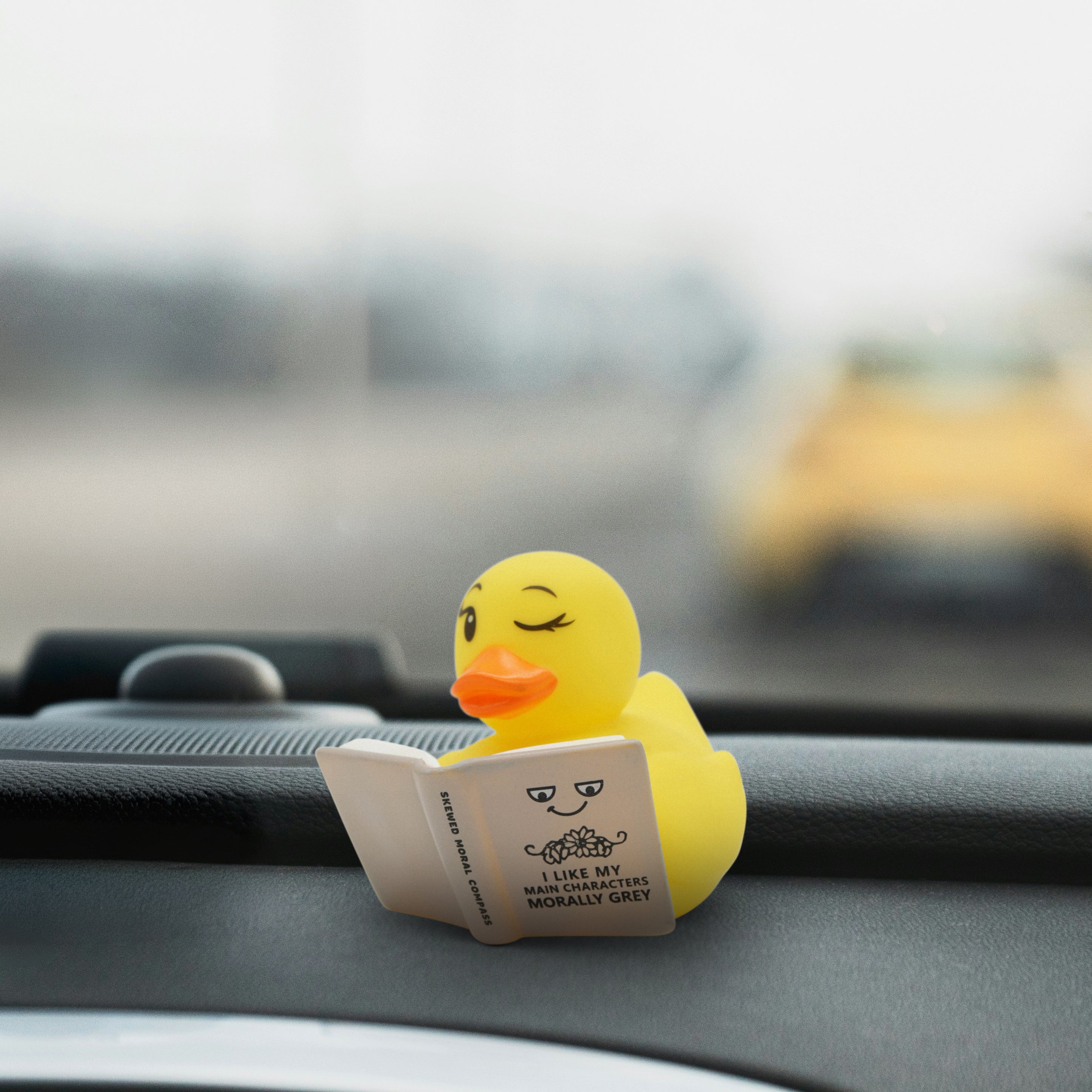 Yellow rubber duck reading a book on a car dashboard with a blurred background