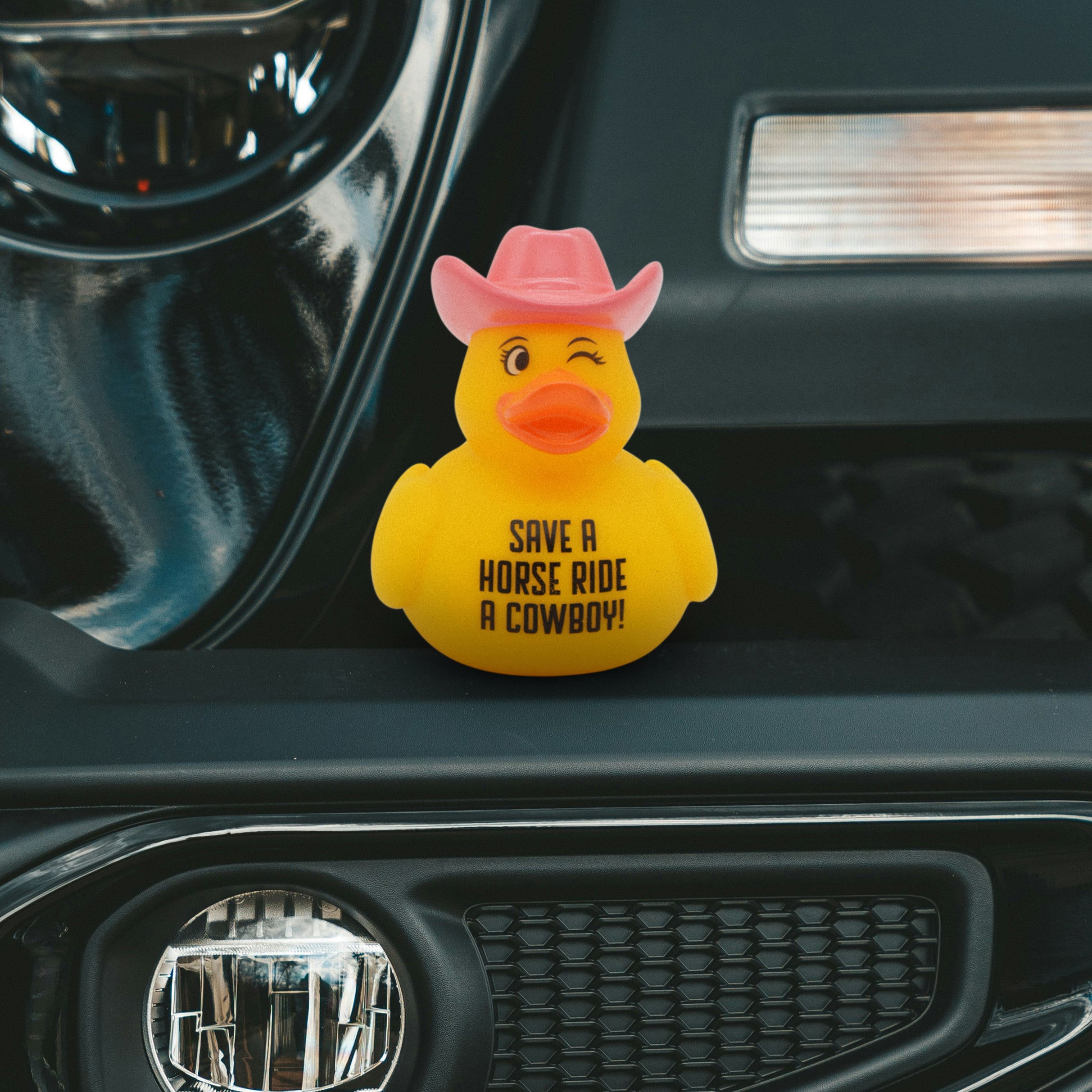 Yellow rubber duck with pink cowboy hat and text 'Save a horse ride a cowboy!' on a car dashboard.