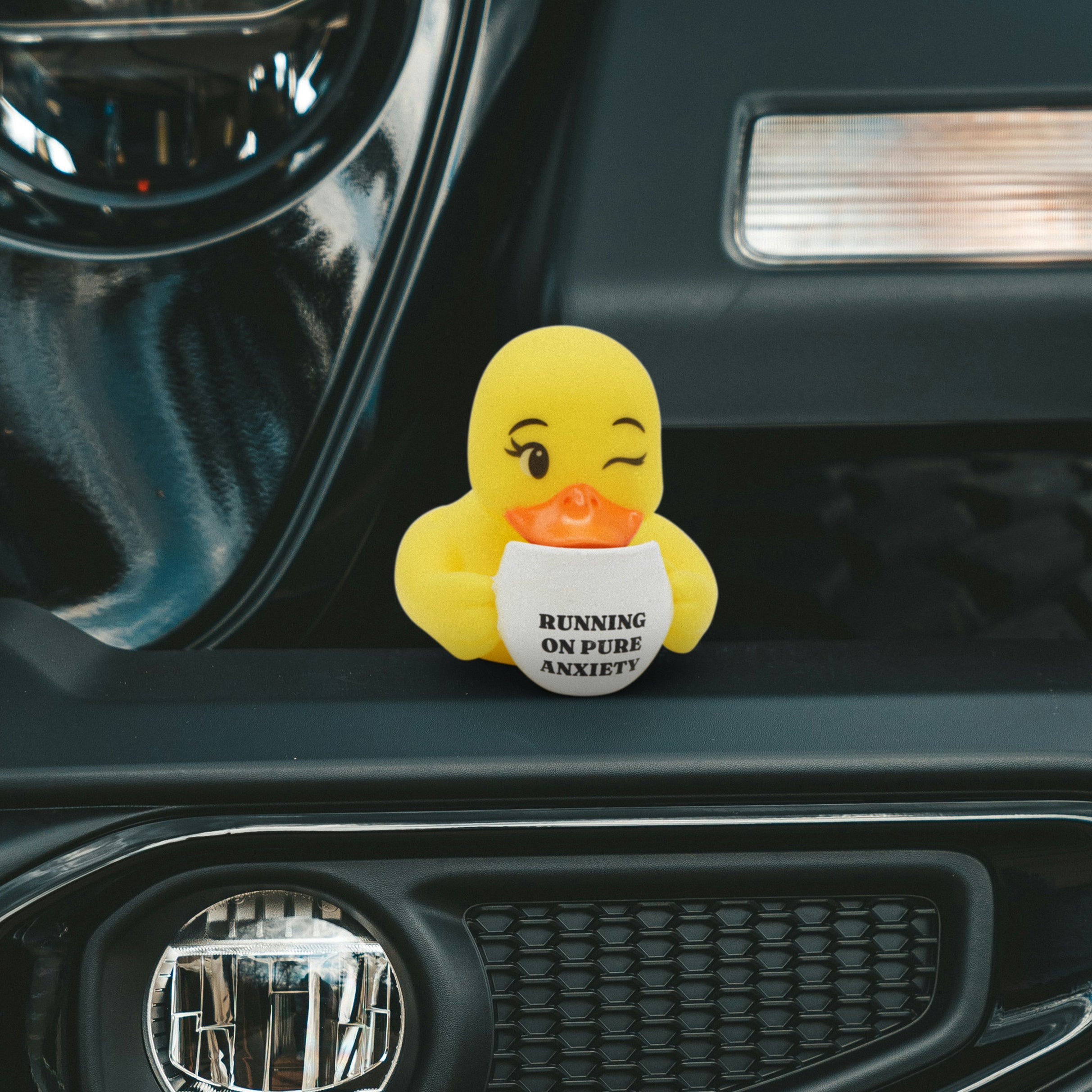 Yellow rubber duck holding a cup that reads 'Running on Pure Anxiety' text on a car dashboard. 