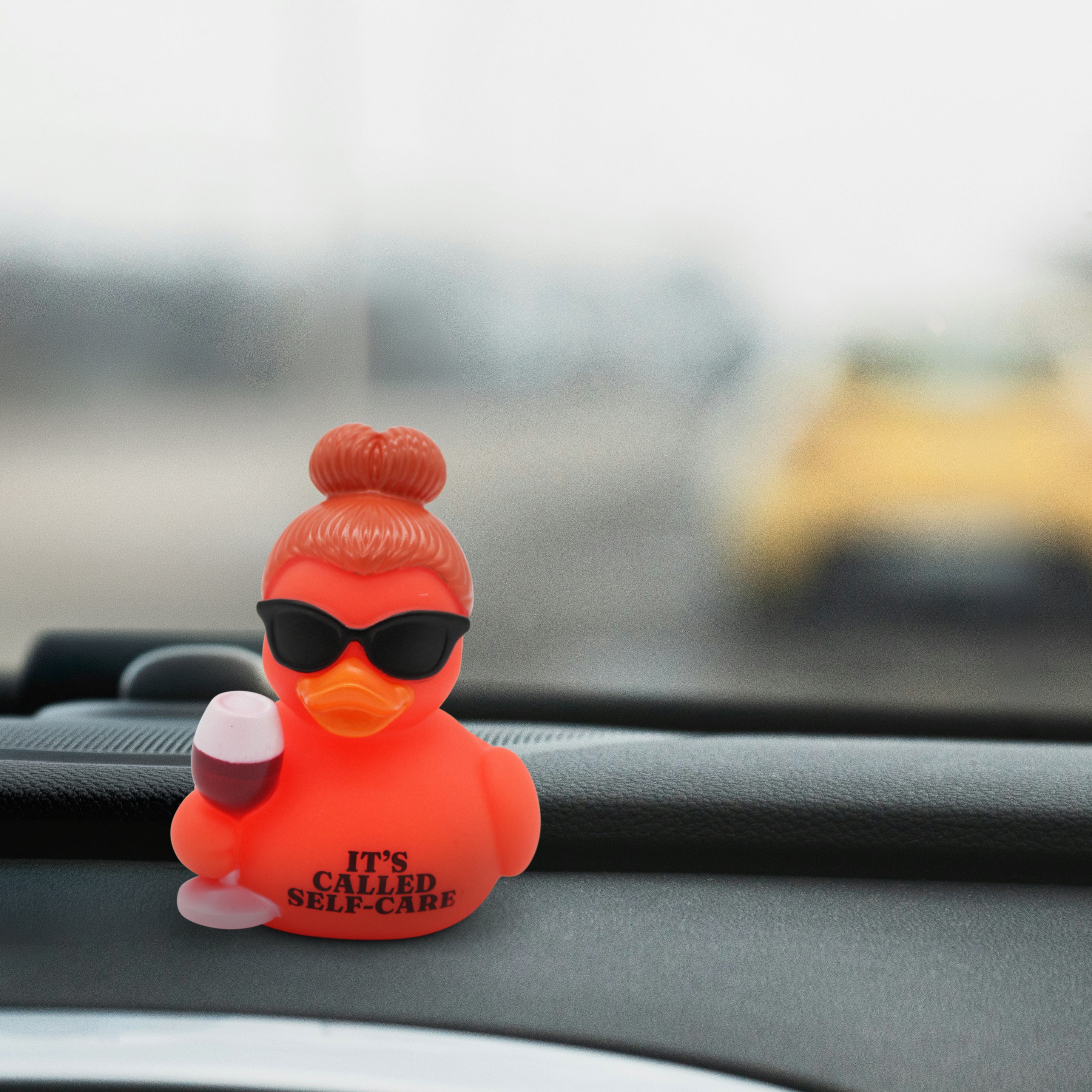 Red rubber duck with sunglasses and a glass of wine with the text 'It's Called Self-Care' on a car dashboard. 