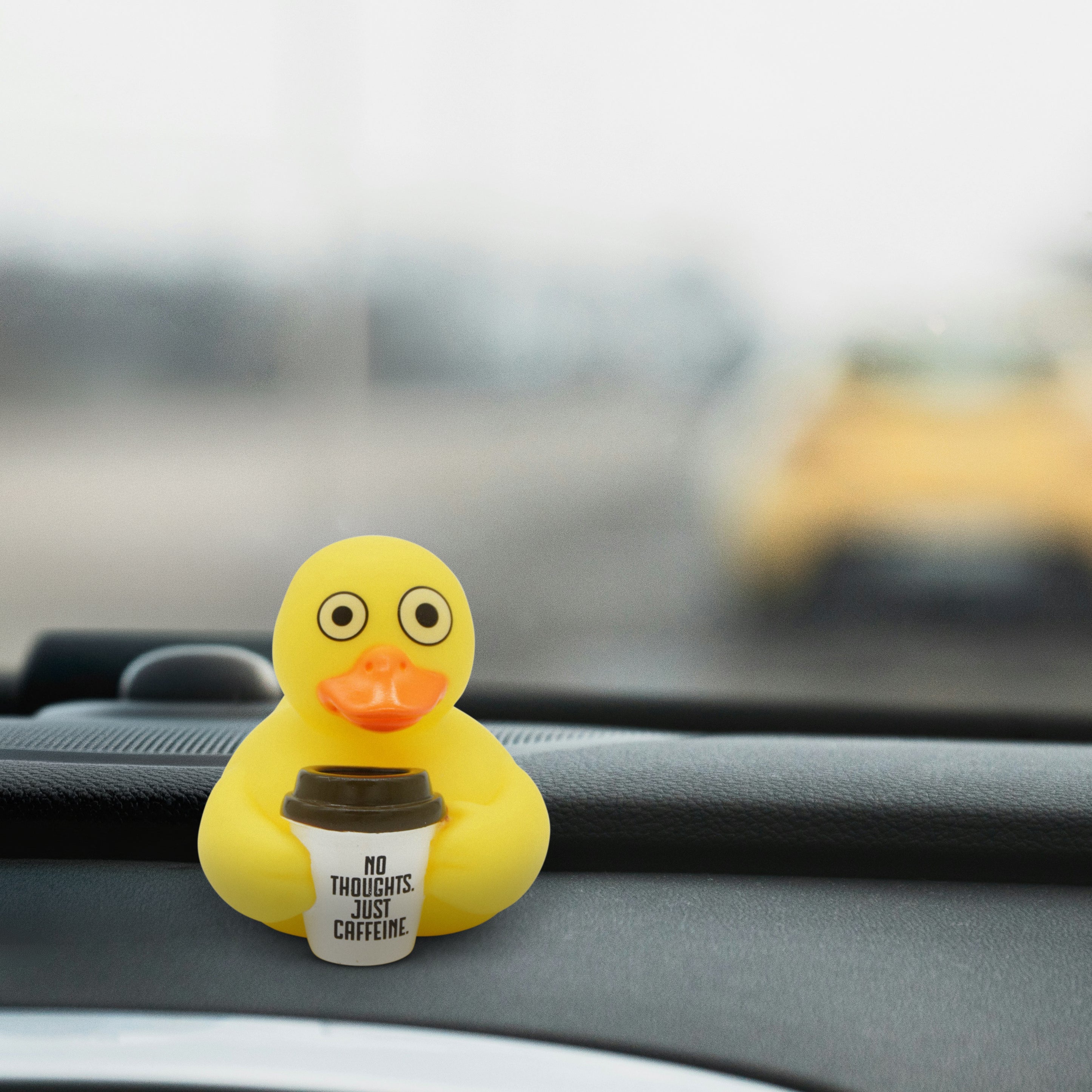 Yellow rubber duck holding a coffee cup with 'No Thoughts. Just Coffee' text on a car dashboard.