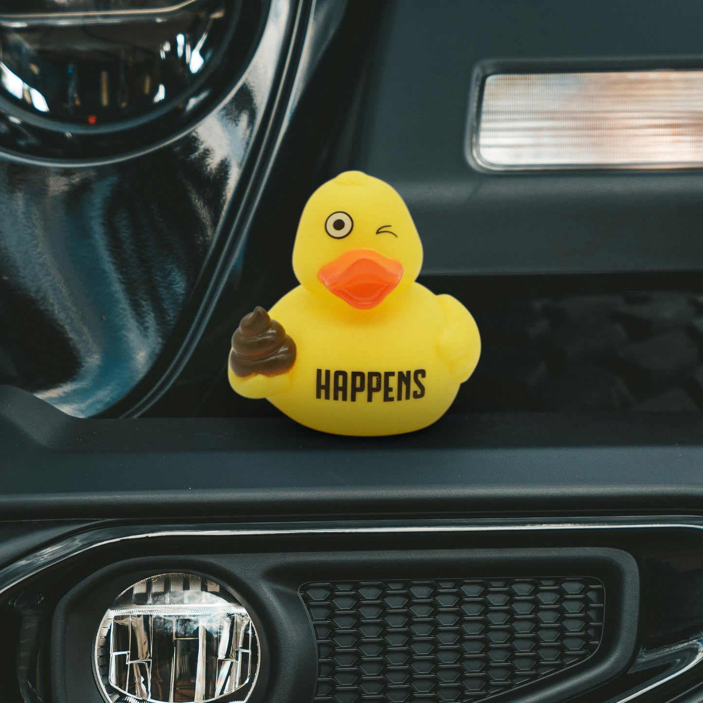 Yellow rubber duck holding a poop with 'HAPPENS' text written on it on a car dashboard