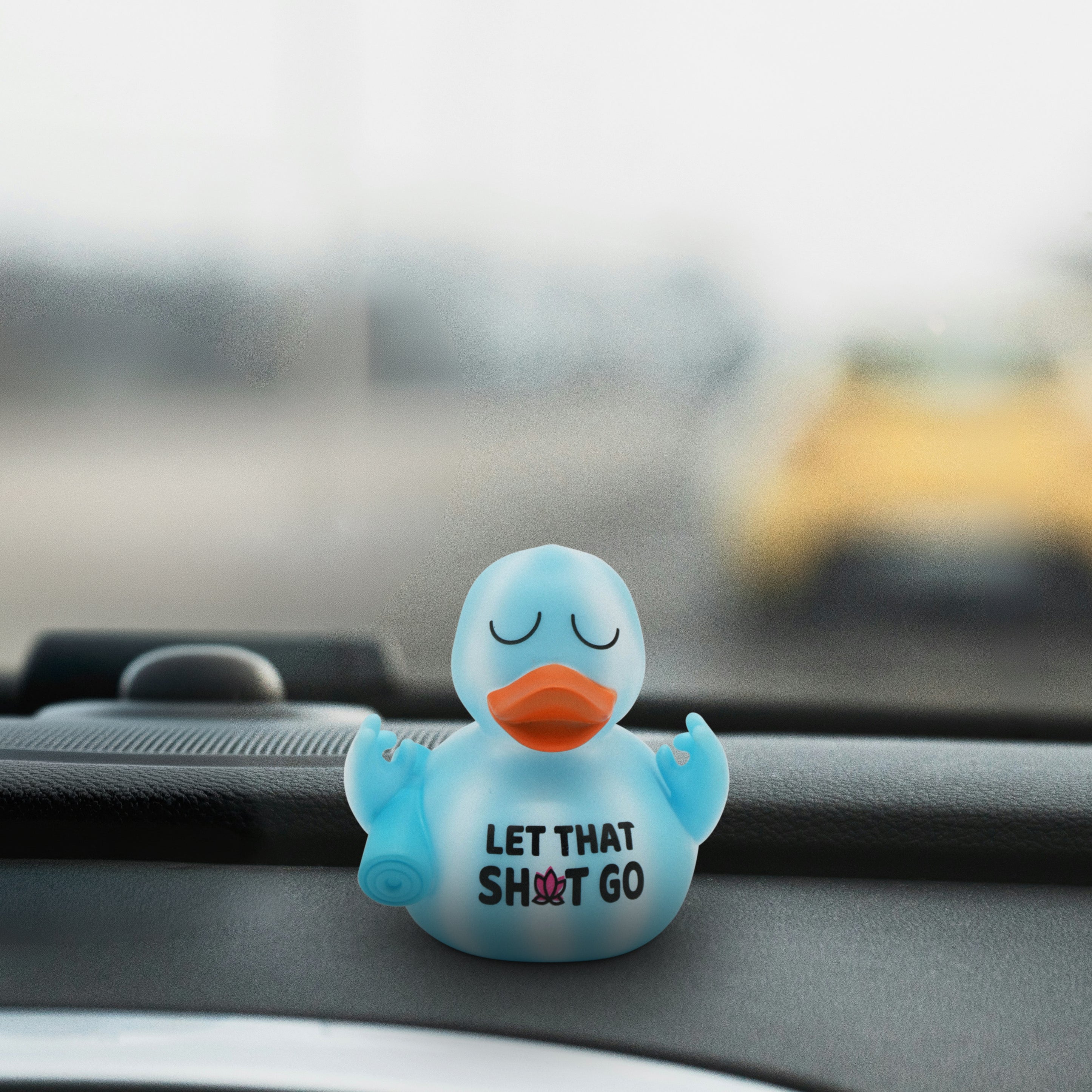 Blue rubber duck holding a yoga mat with 'Let That Sh*t Go' text on a car dashboard