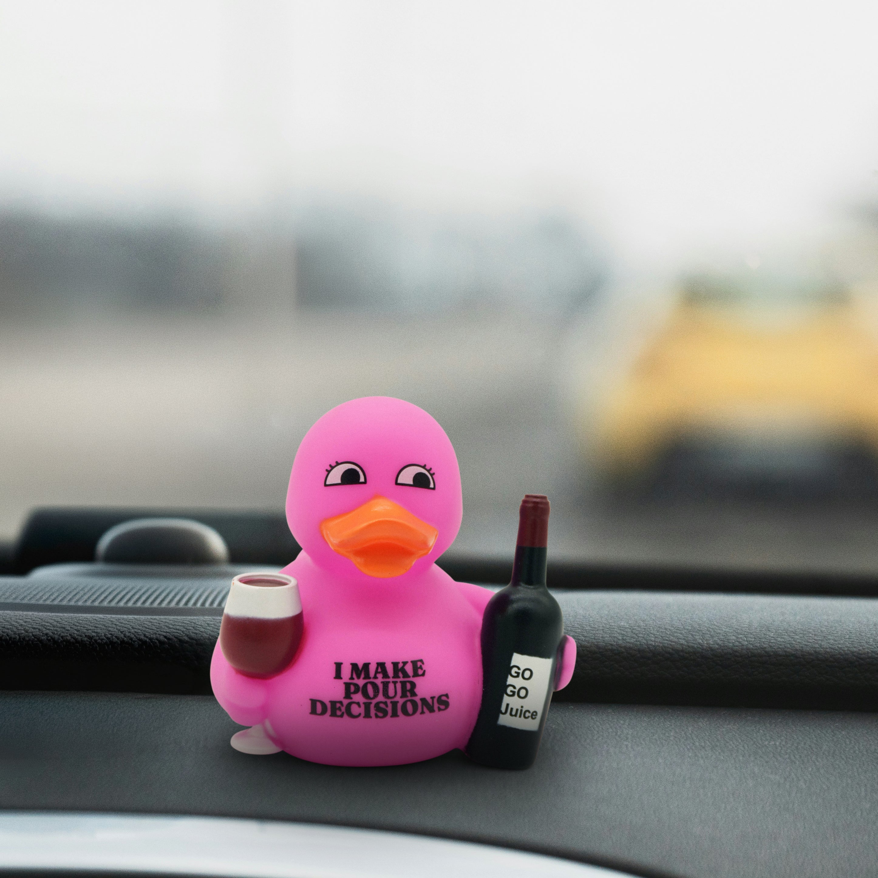 Pink rubber duck that reads "I make pour decisions" holding a bottle and cup on a car dashboard.