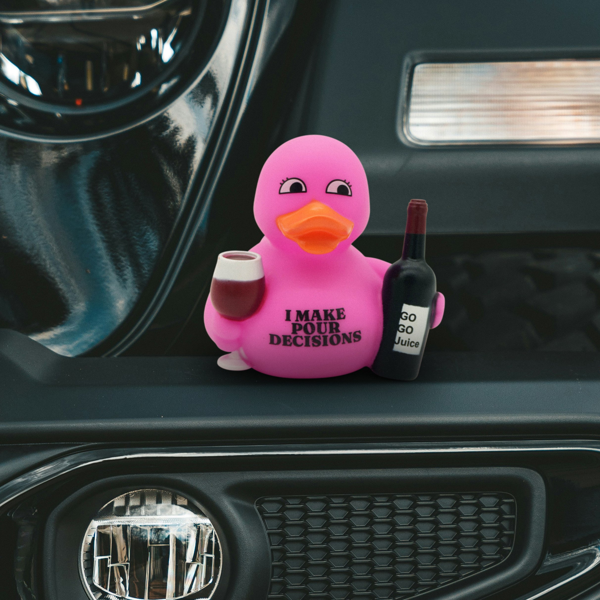 Pink rubber duck that reads "I make pour decisions" holding a glass of wine and a bottle on a car dashboard.