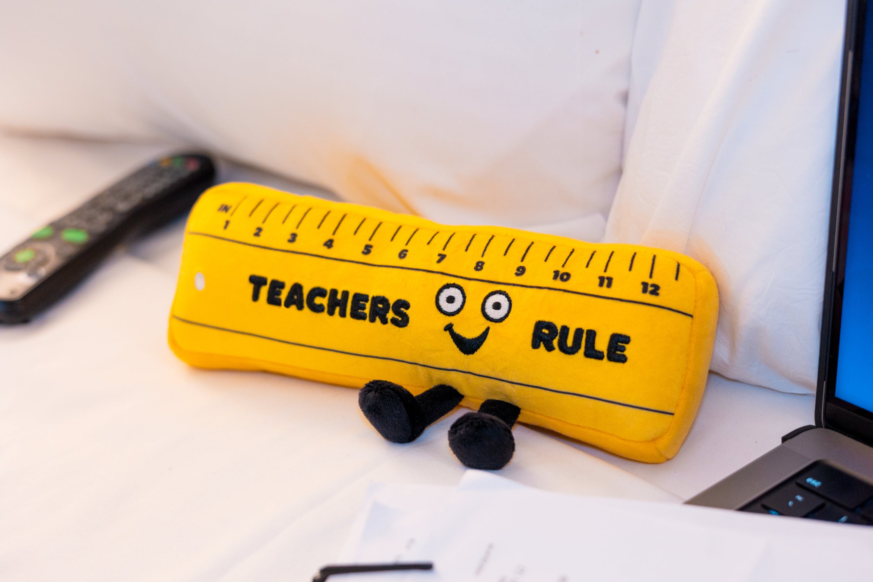 A plush shaped like a yellow ruler with a face and feet, featuring the text 'TEACHERS RULE' placed on a bed.