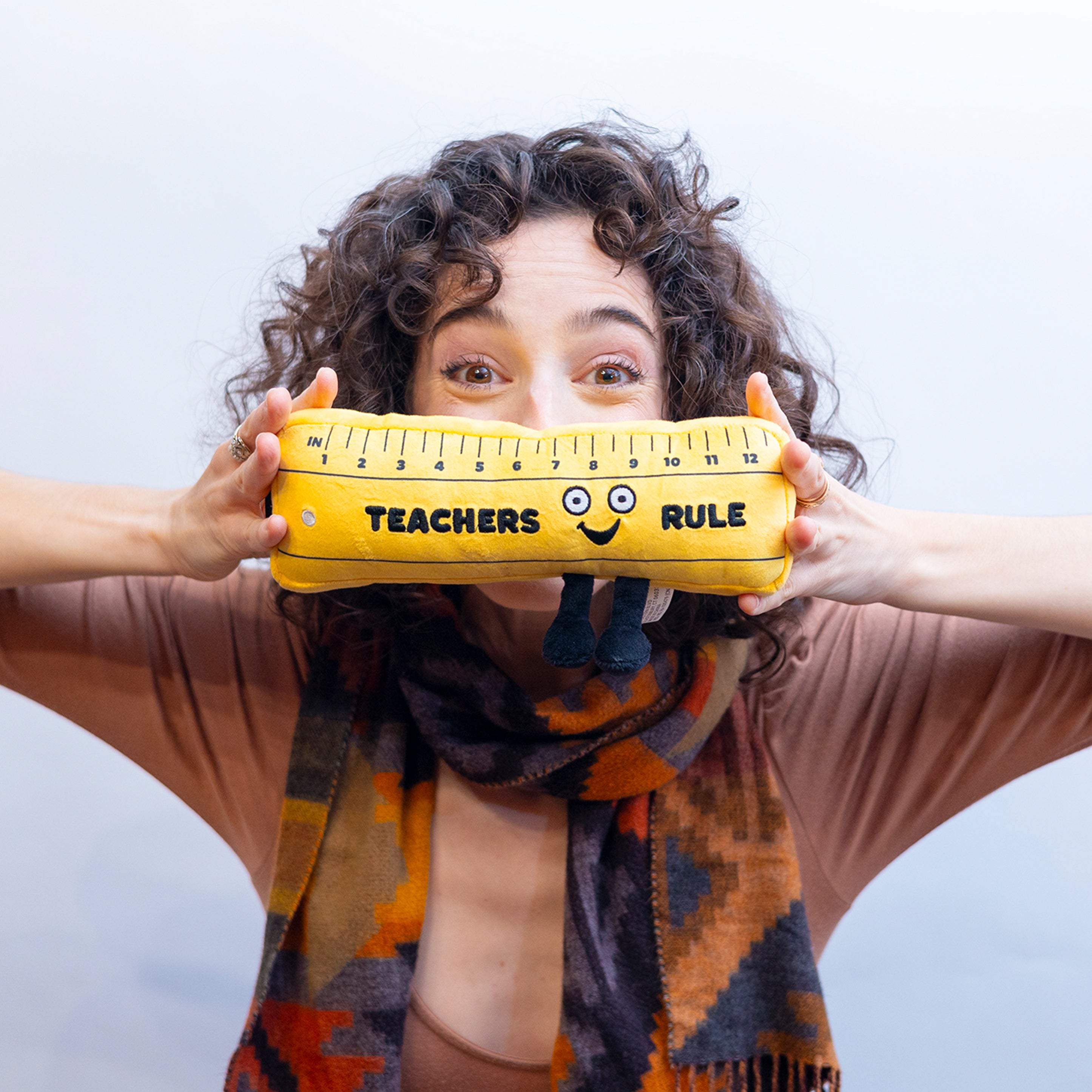 A woman holding a plush shaped like a yellow ruler with a face and feet, featuring the text 'TEACHERS RULE' on the top.