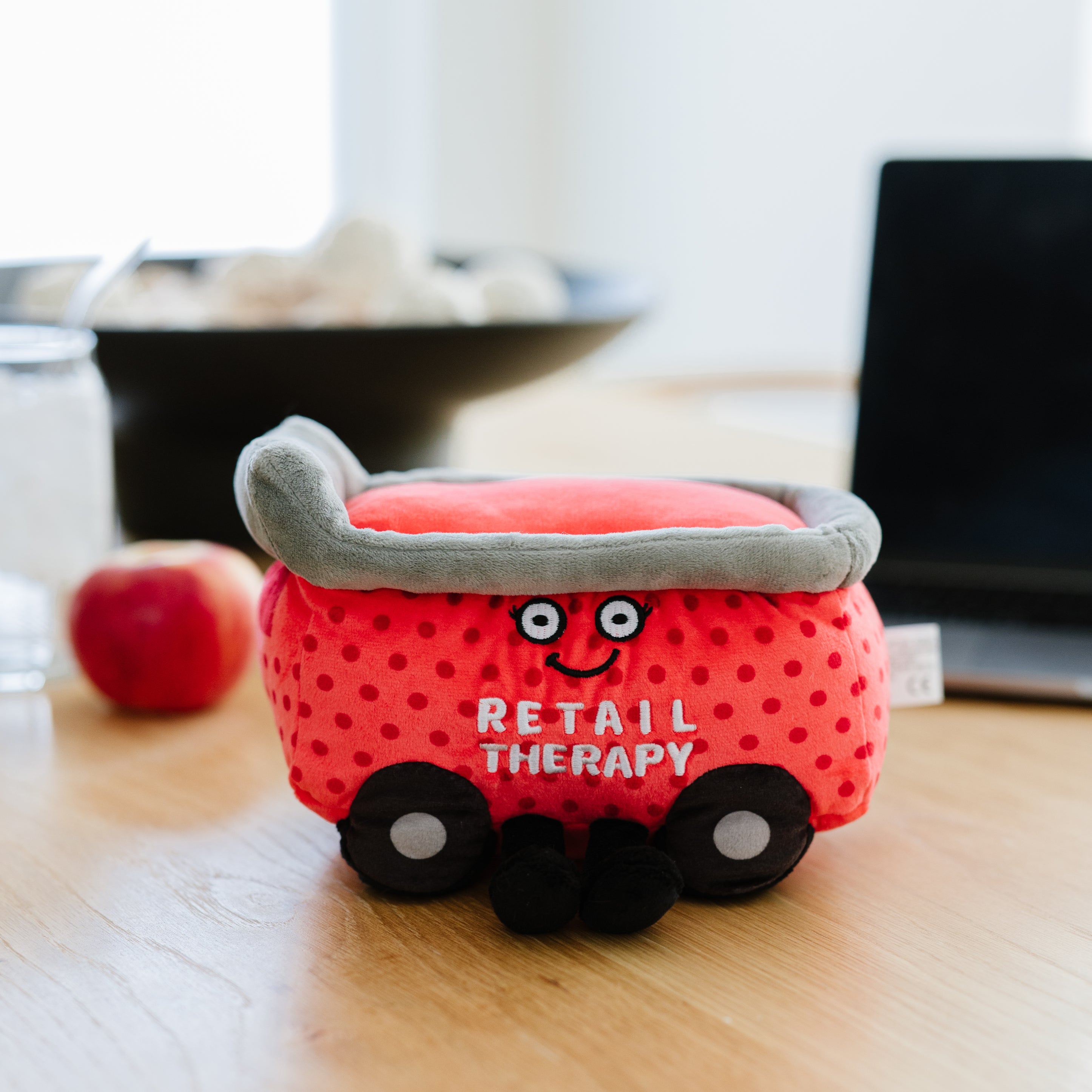 A plush resembling a red shopping cart with the text 'RETAIL THERAPY' written on it placed on a kitchen table. 