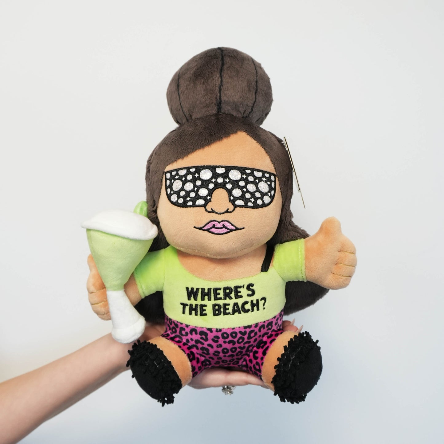 A hand holding a plush made to look like Snooki. She's got her signature poof, is wearing bejeweled sunglasses, and is wearing a shirt that says 'Where's the Beach?'
