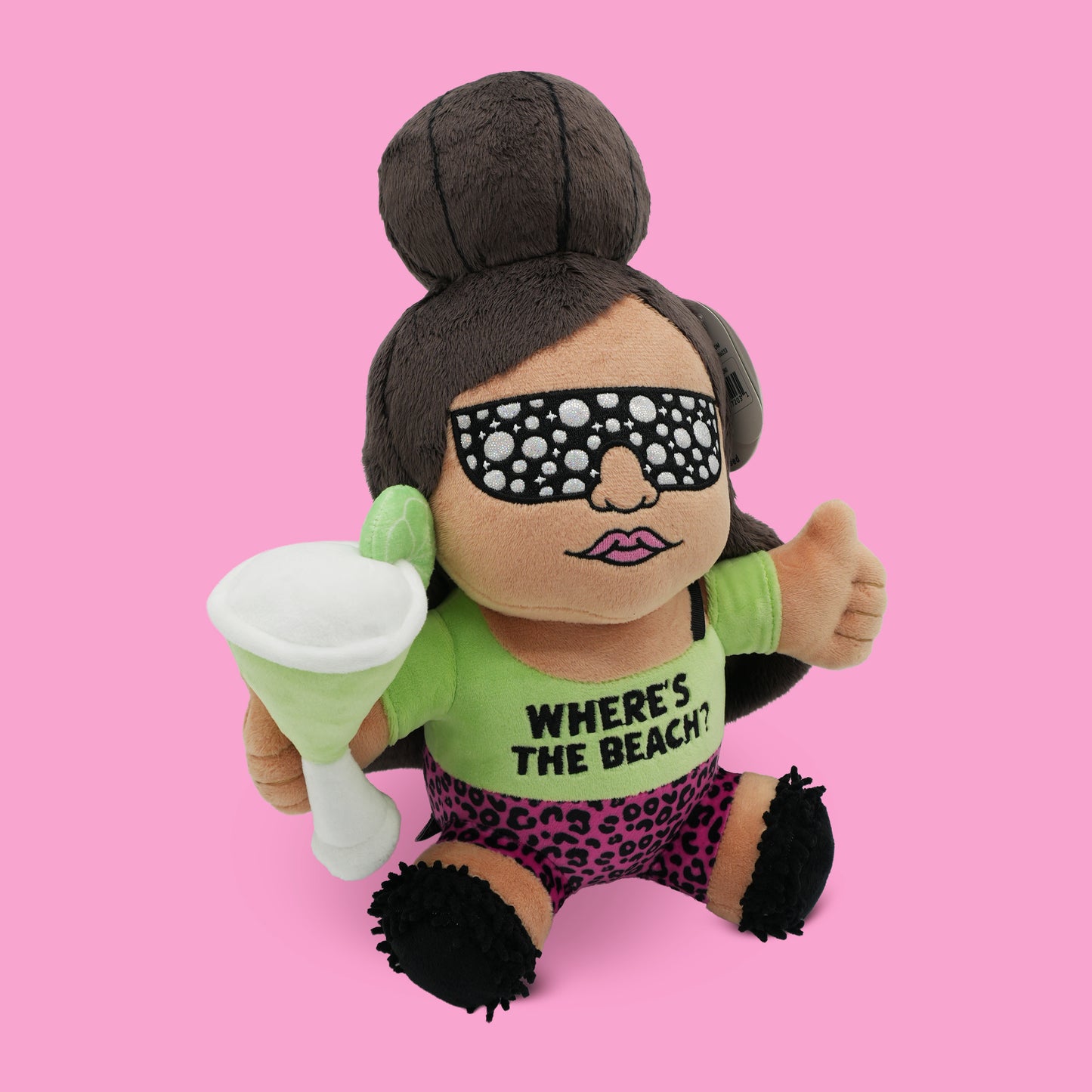A plush made to look like Snooki. She's got her signature poof, is wearing bejeweled sunglasses, is holding a martini glass, and is wearing a shirt that says 'Where's the Beach?' placed on a pink background.