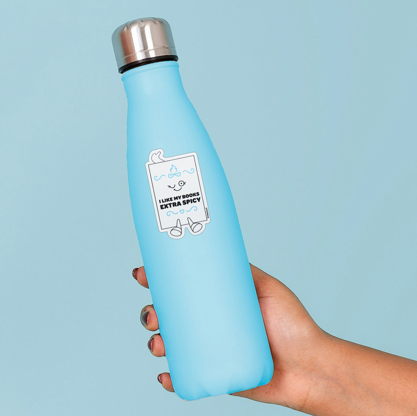 A light blue insulated water bottle with a stainless steel cap, held in a hand against a solid light blue background. The bottle features a white graphic label that reads 'I LIKE MY BOOKS EXTRA SPICY' with cartoon graphics.