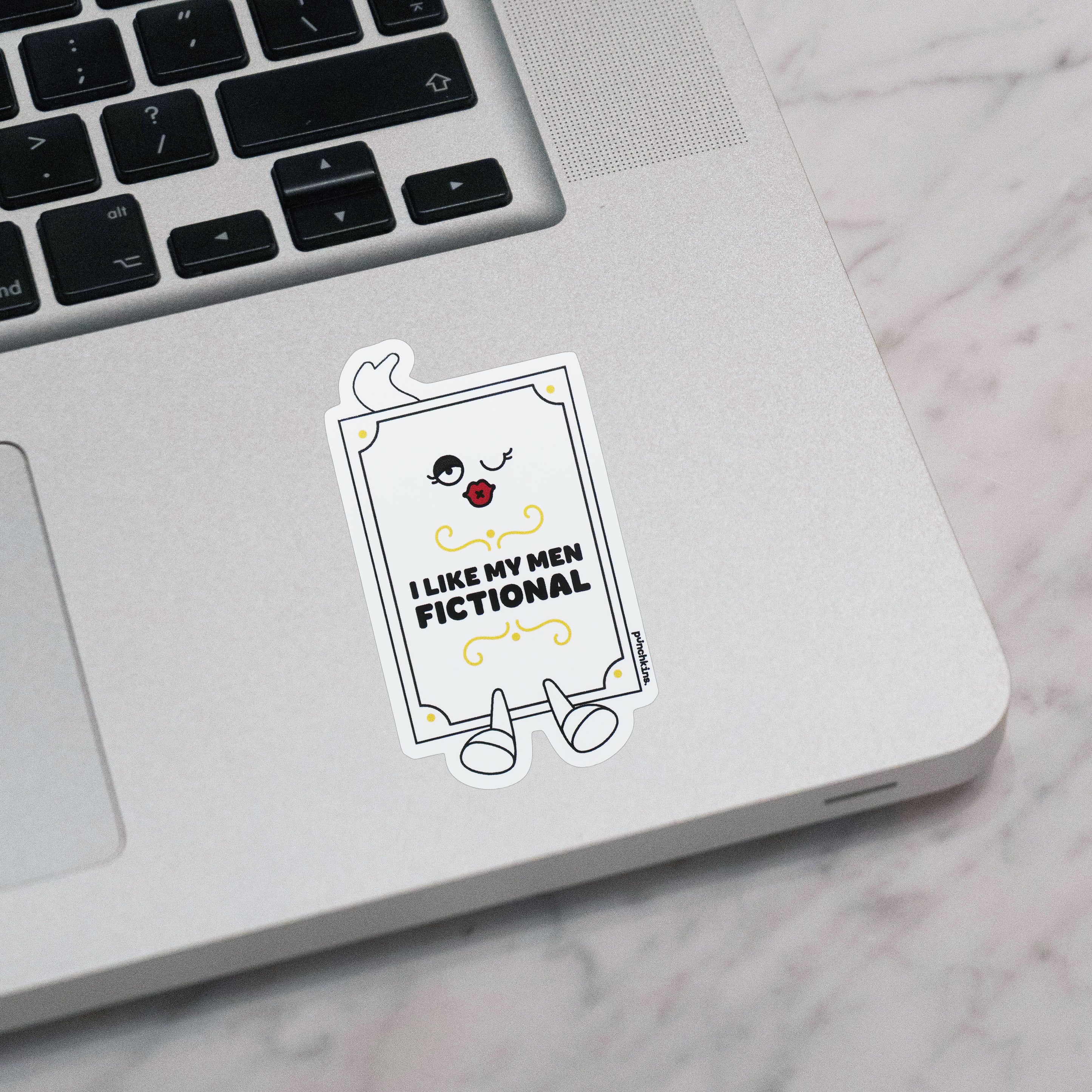A die-cut sticker with a cartoon book character and the text 'I LIKE MY MEN FICTIONAL' applied to a silver laptop on a white marble surface.