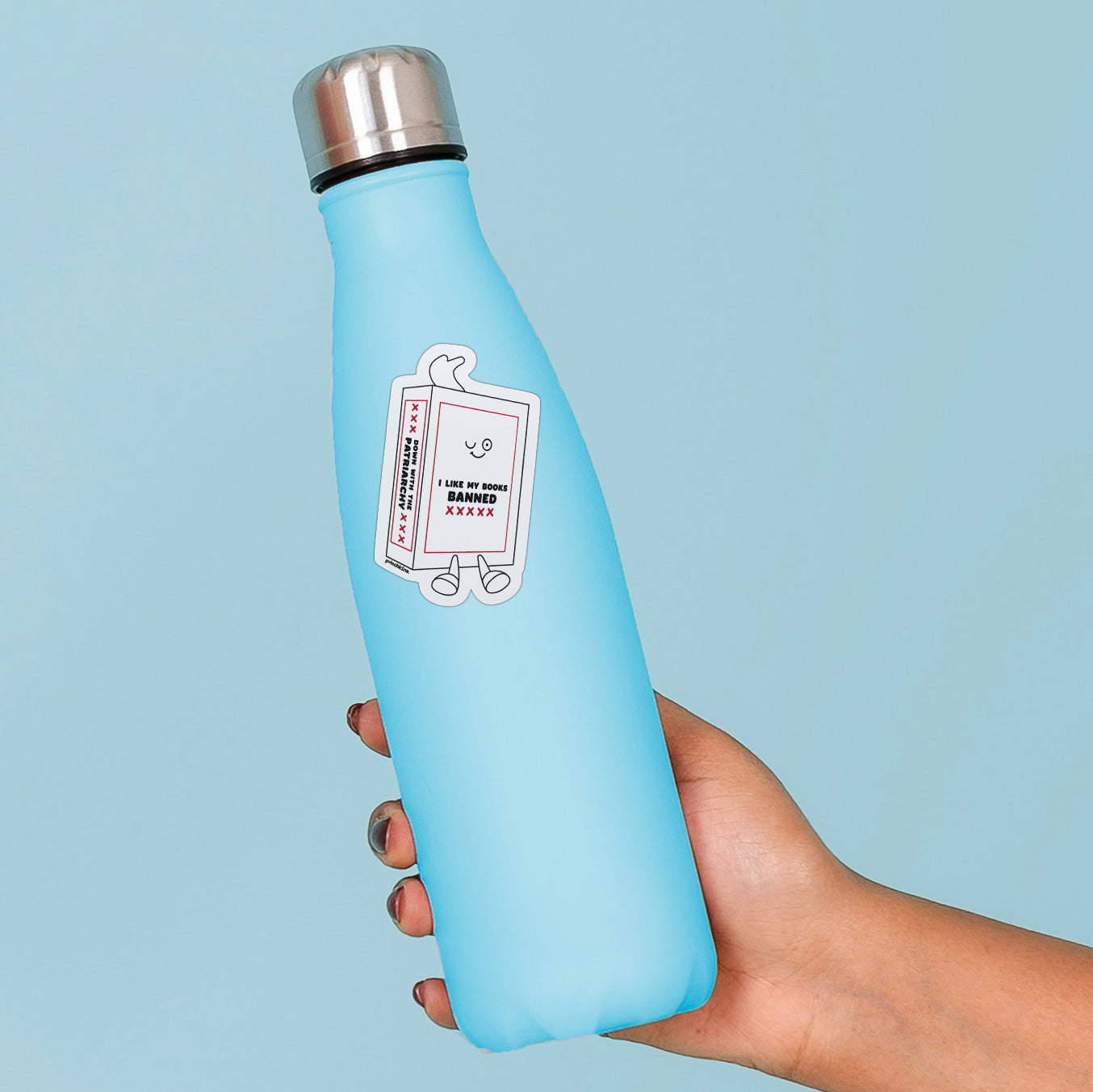 A light blue reusable water bottle with a stainless steel cap, held in a hand against a solid light blue background. The bottle features a cartoon sticker of a book with the text 'I LIKE MY BOOKS BANNED.'
