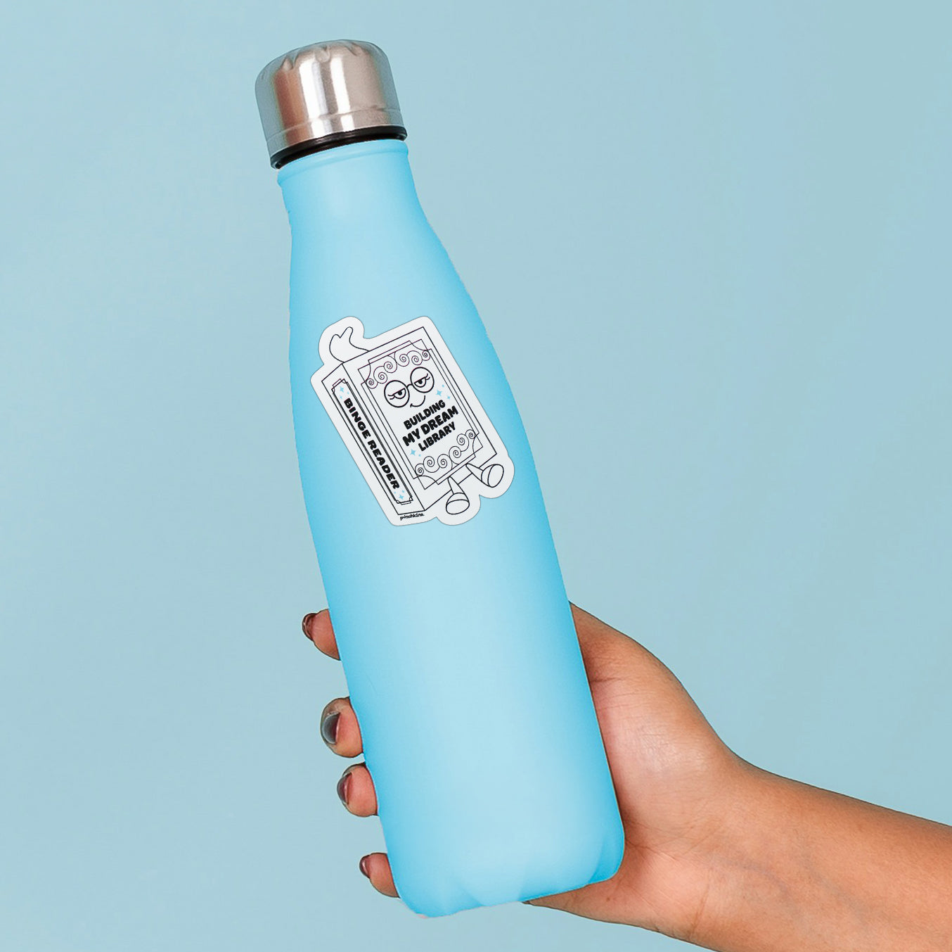 A light blue insulated water bottle with a stainless steel cap, held in a hand against a solid light blue background. The bottle features a white sticker on the front with a cartoon book with the text 'BUILDING MY DREAM LIBRARY' on the cover.