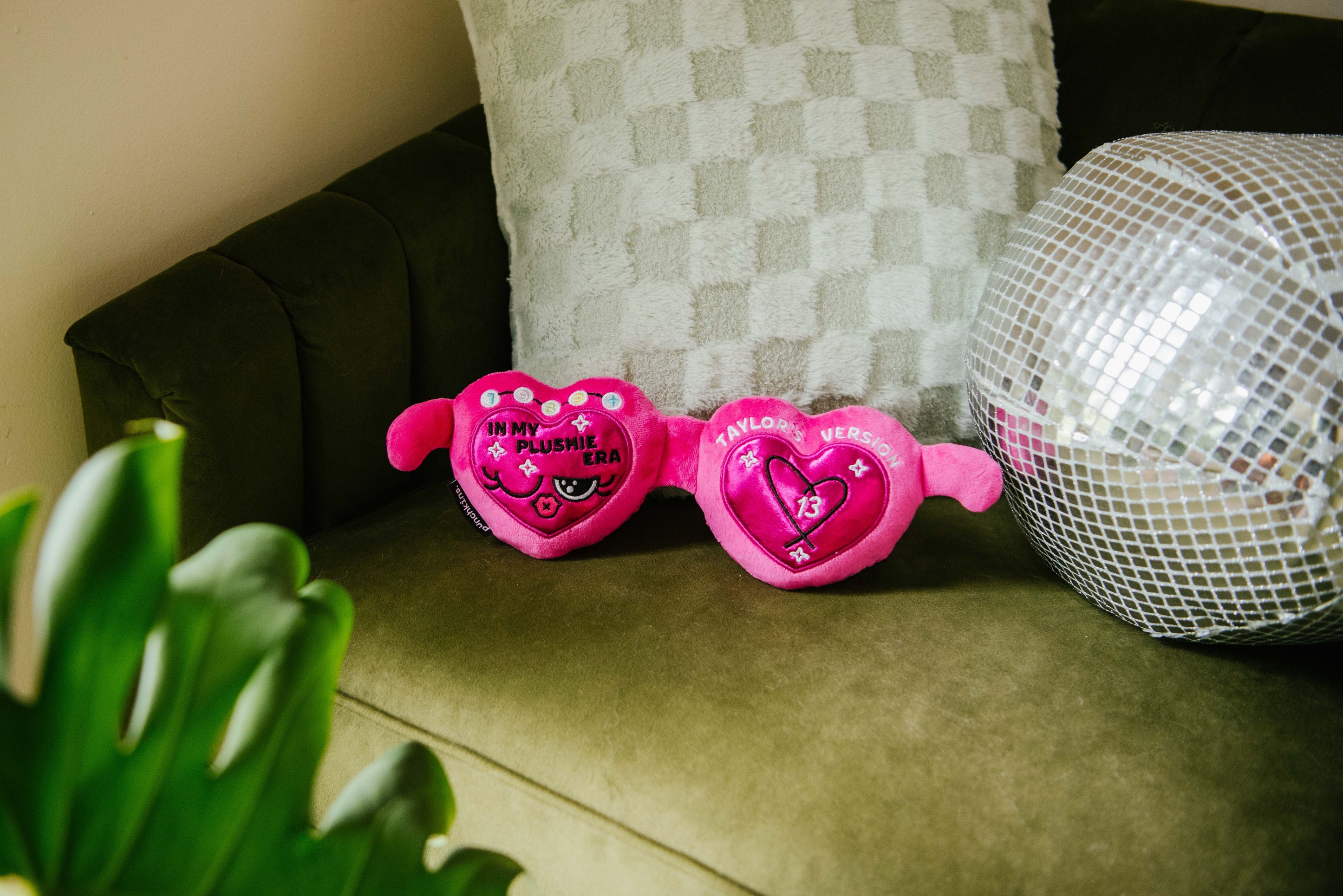 A plush resembling pink sunglasses with hearts and text that reads, "In my plushie era" placed on a couch.