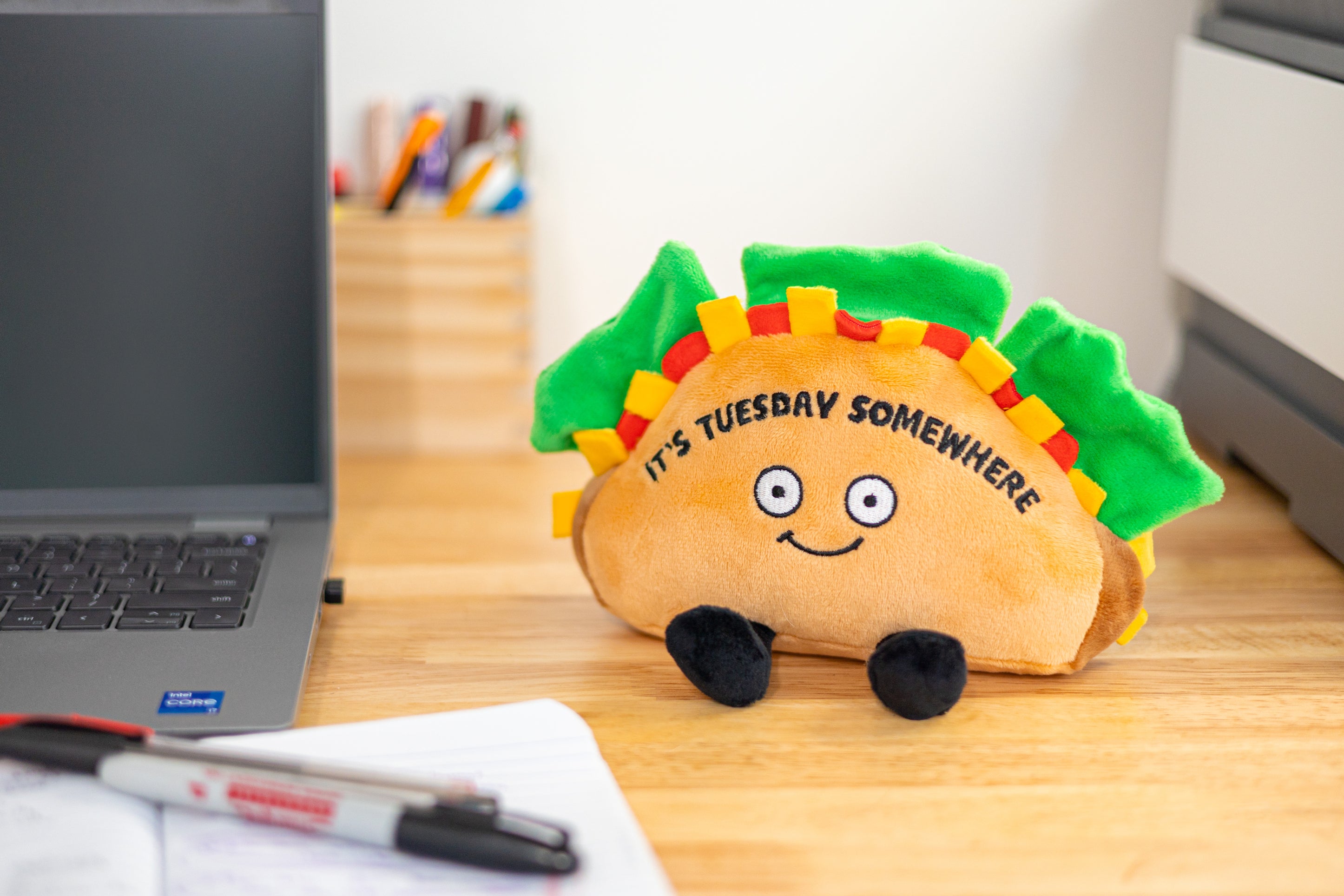 A plush shaped like a taco with a smiling face, green lettuce leaves, and the text 'IT'S TUESDAY SOMEWHERE' on the shell placed on a desk.