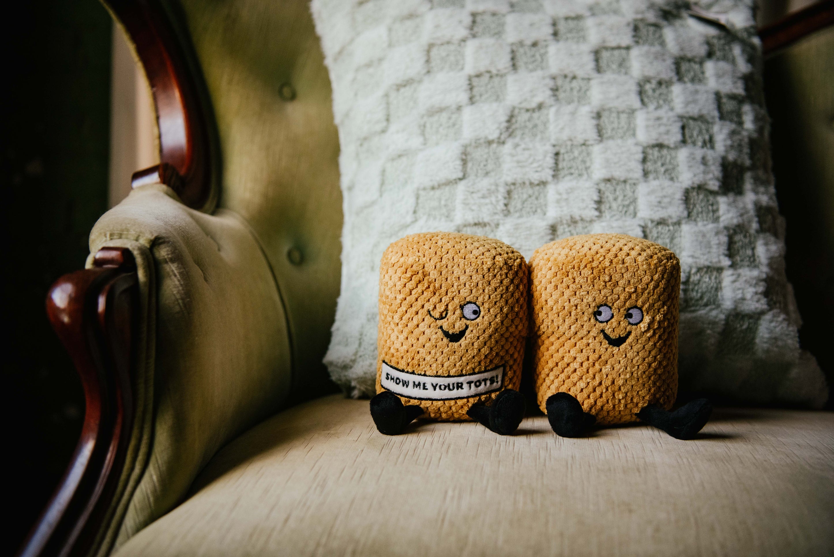 A plushie designed to resemble a pair of tater tots with faces and text that reads 'SHOW ME YOUR TOTS!' placed on a couch.