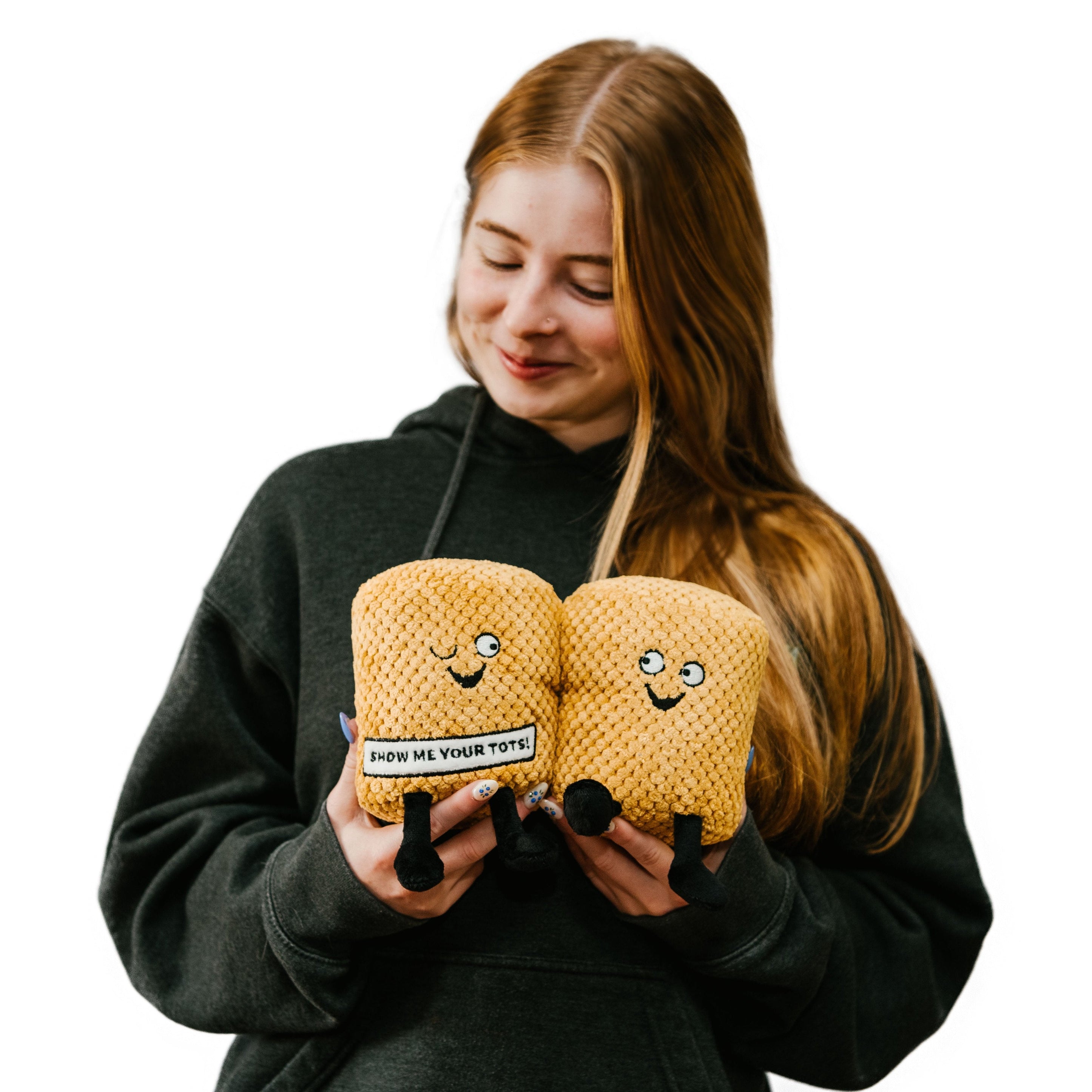 A woman holding a plushie designed to resemble a pair of tater tots with faces and text that reads 'SHOW ME YOUR TOTS!'