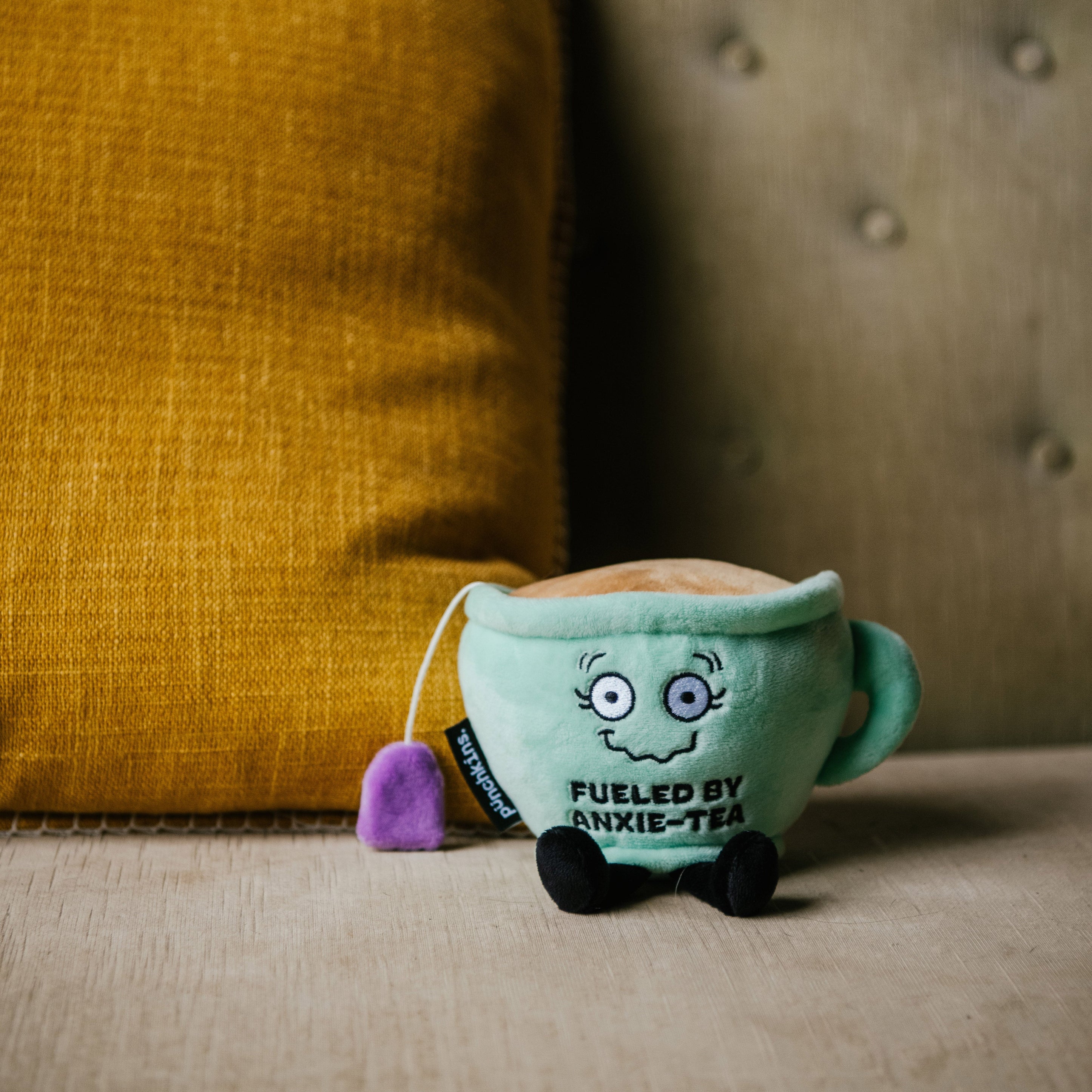 A plush designed to look like a teacup with a face and text that reads 'FUELED BY ANXIE-TEA' placed on a couch.