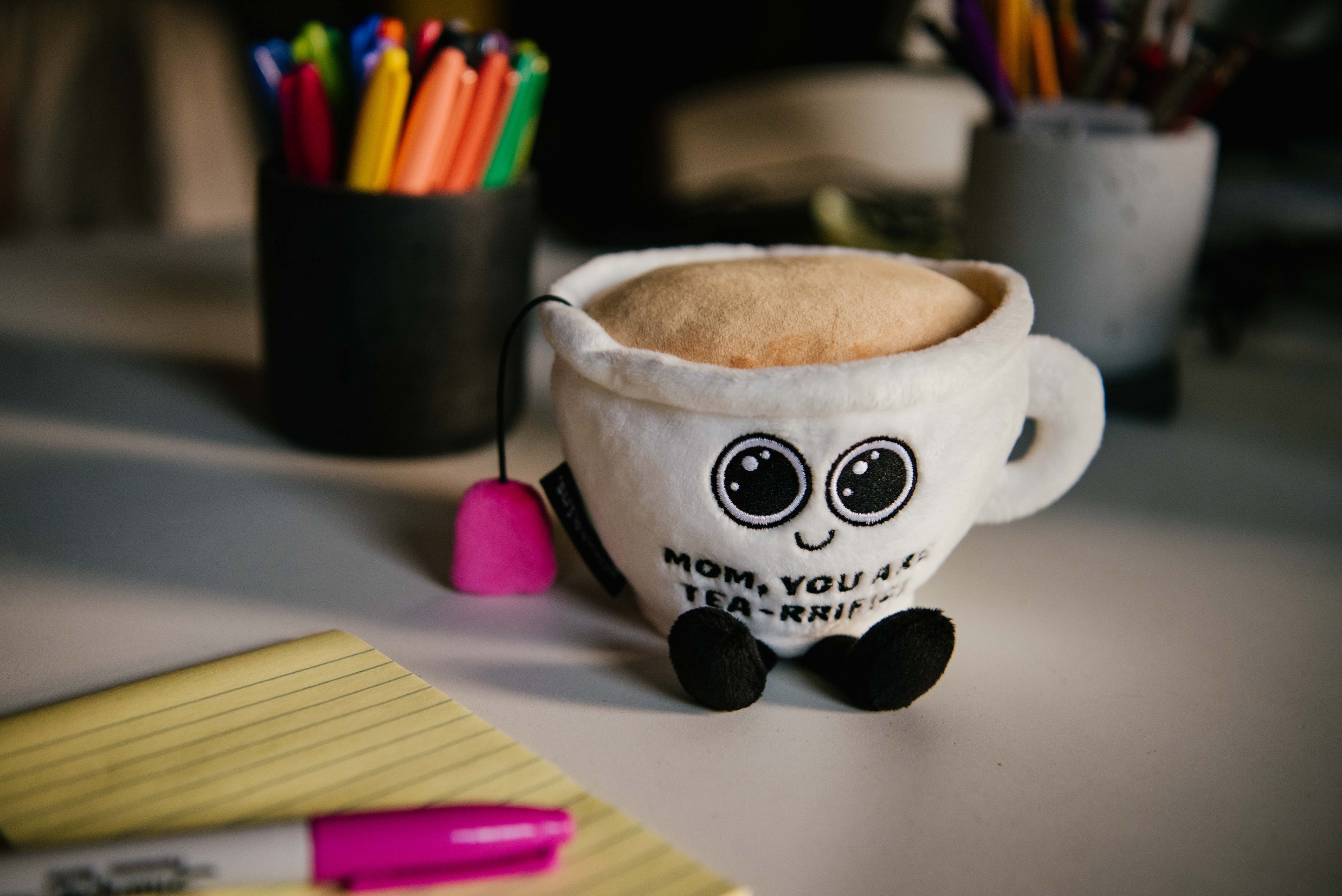 A plush shaped like a teacup with a face, eyes, a teabag hanging from its side, and the text 'MOM, YOU ARE TEA-RRIFIC' is visible on it placed on a desk.