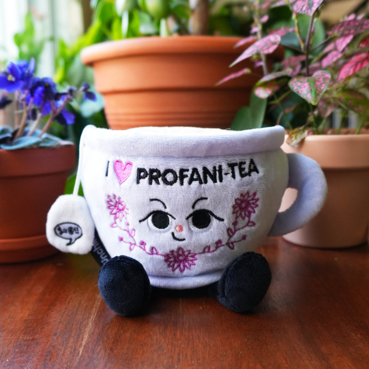 Plush toy resembling a teacup with 'I love PROFANI-TEA' text, surrounded by potted plants on a wooden surface.