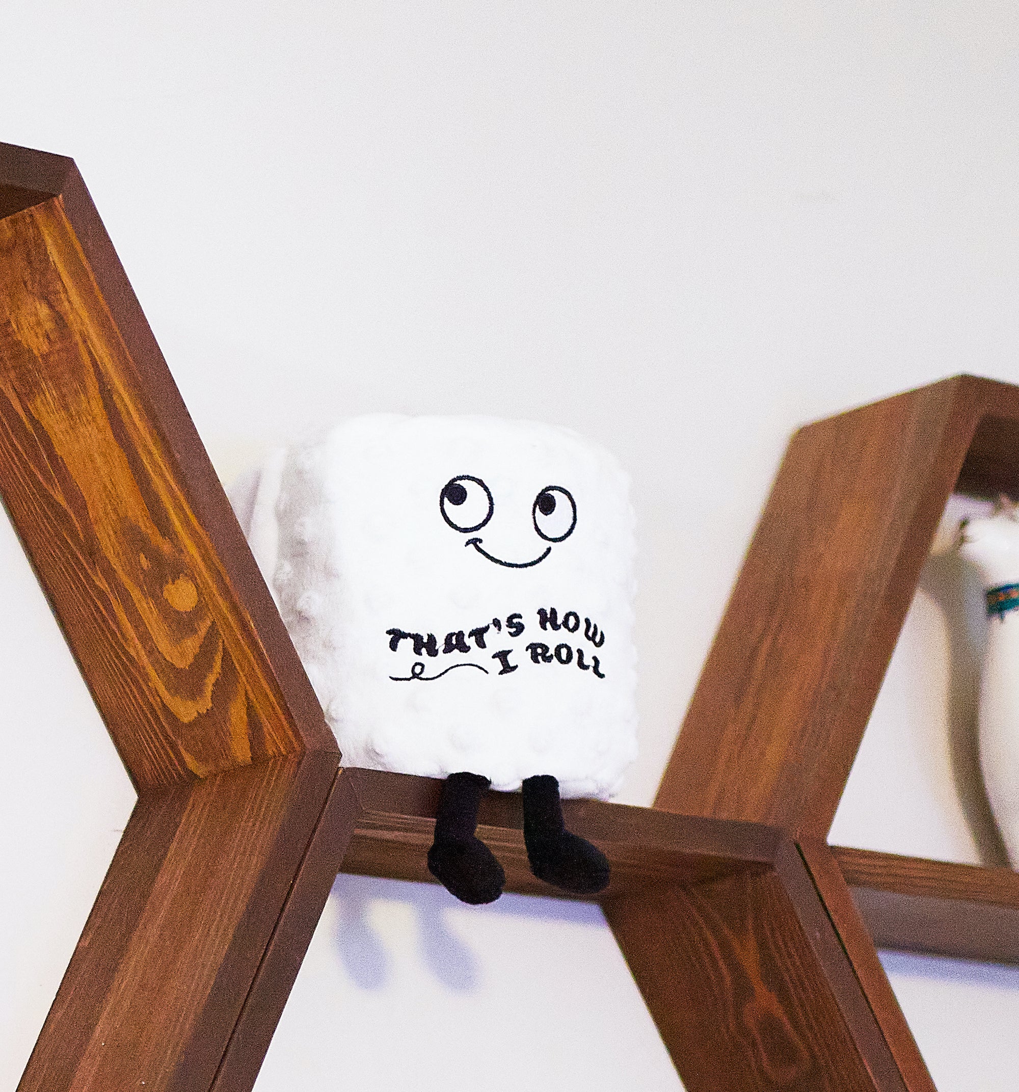 A plush toilet paper roll with a smiling face and the phrase 'That's How I Roll' printed on it placed on a shelf.