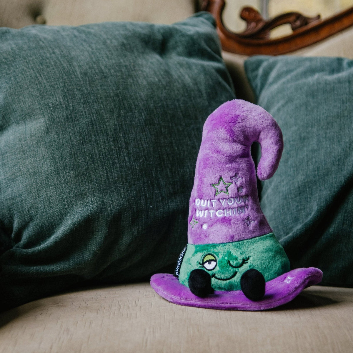 A plush witch hat featuring text that reads, "quit your witching'" placed on a couch.