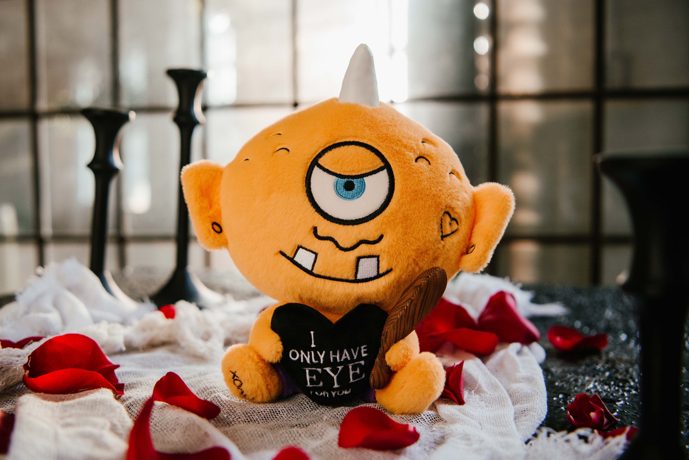 A cyclops plush toy that reads, "I only have eye for you," sitting on a bed of rose petals. 
