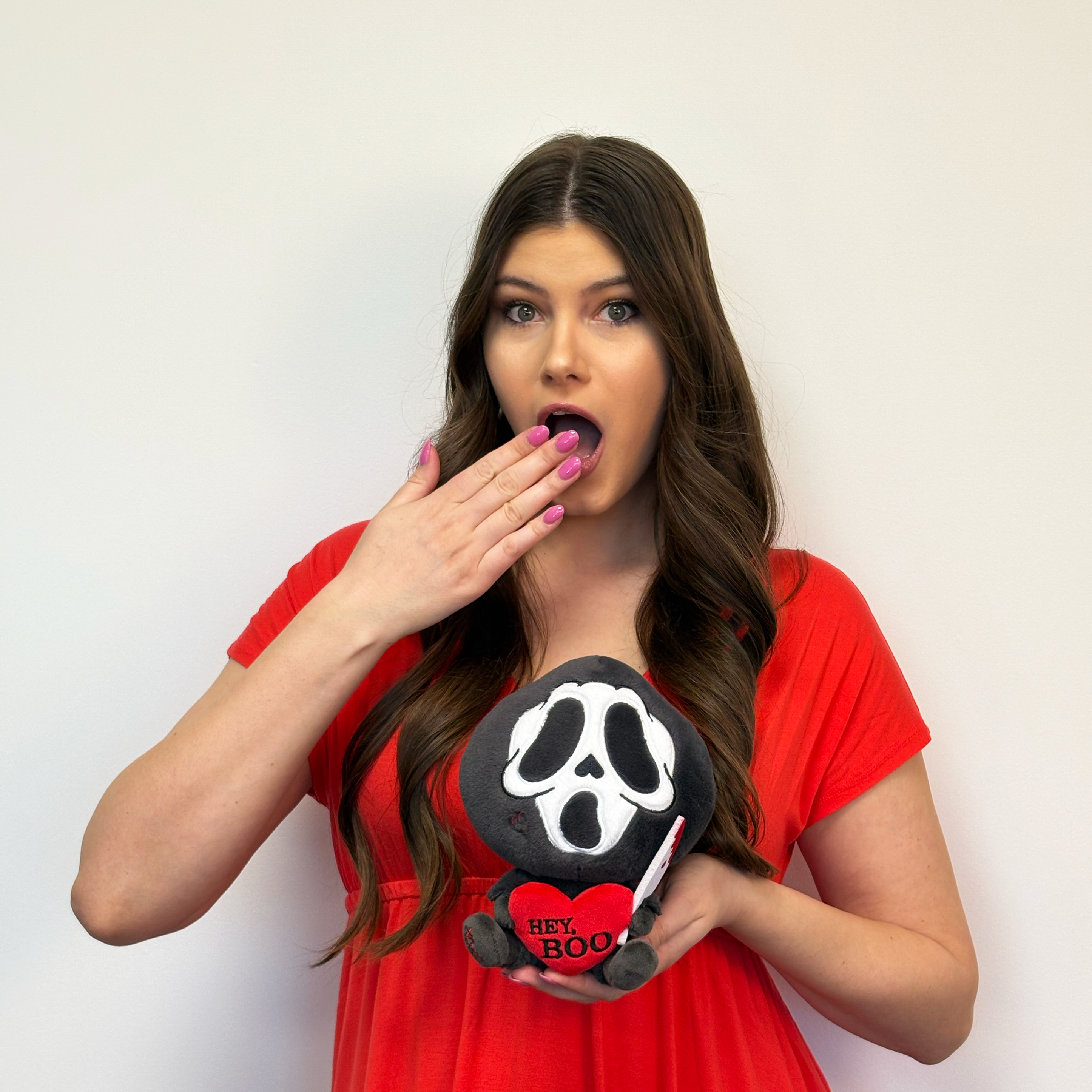 Brunette woman holding ghost face killer plush that says, "Hey, boo," in a heart. 