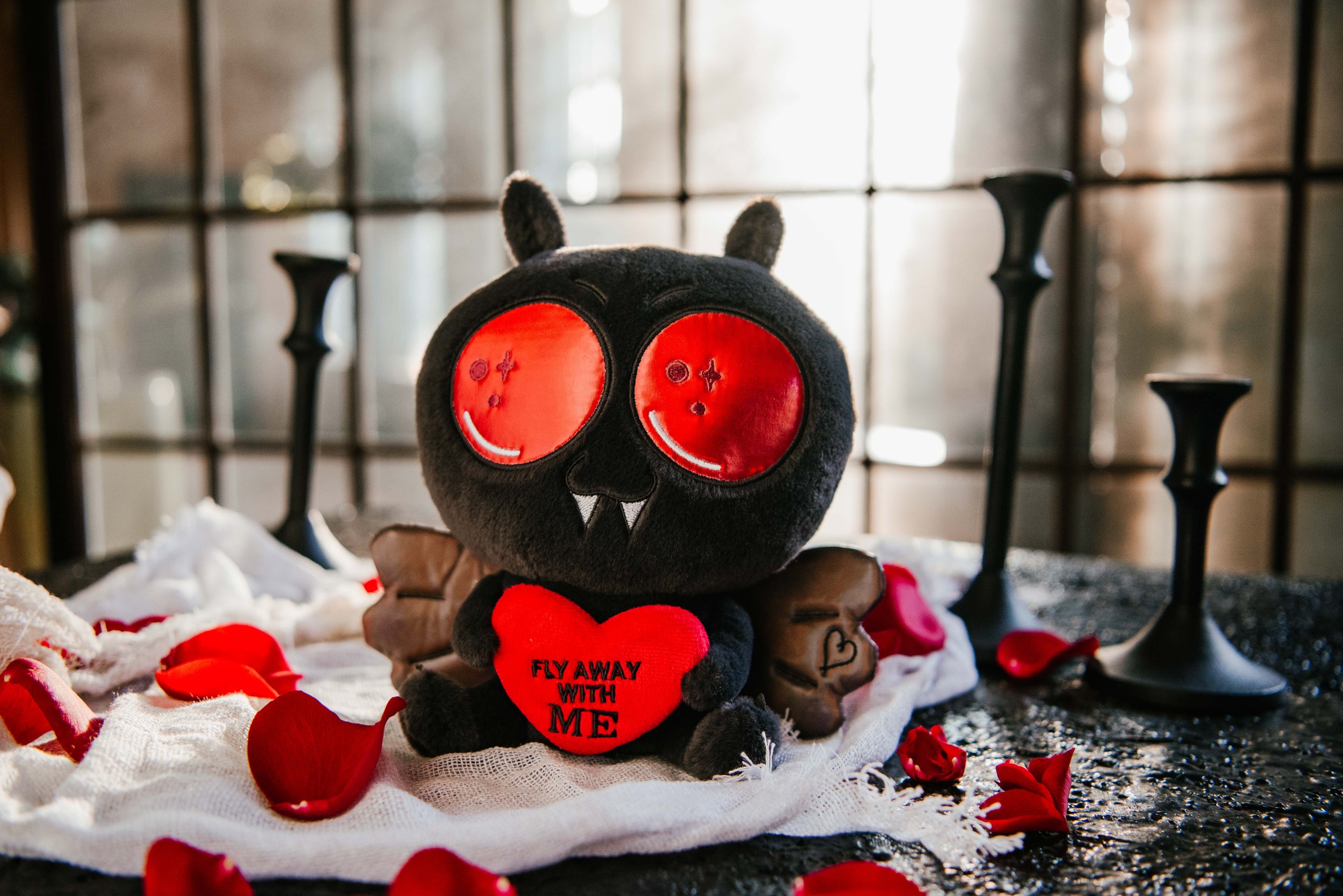 Mothman plushie that reads, "Fly away with me," sitting on a bed of rose petals. 