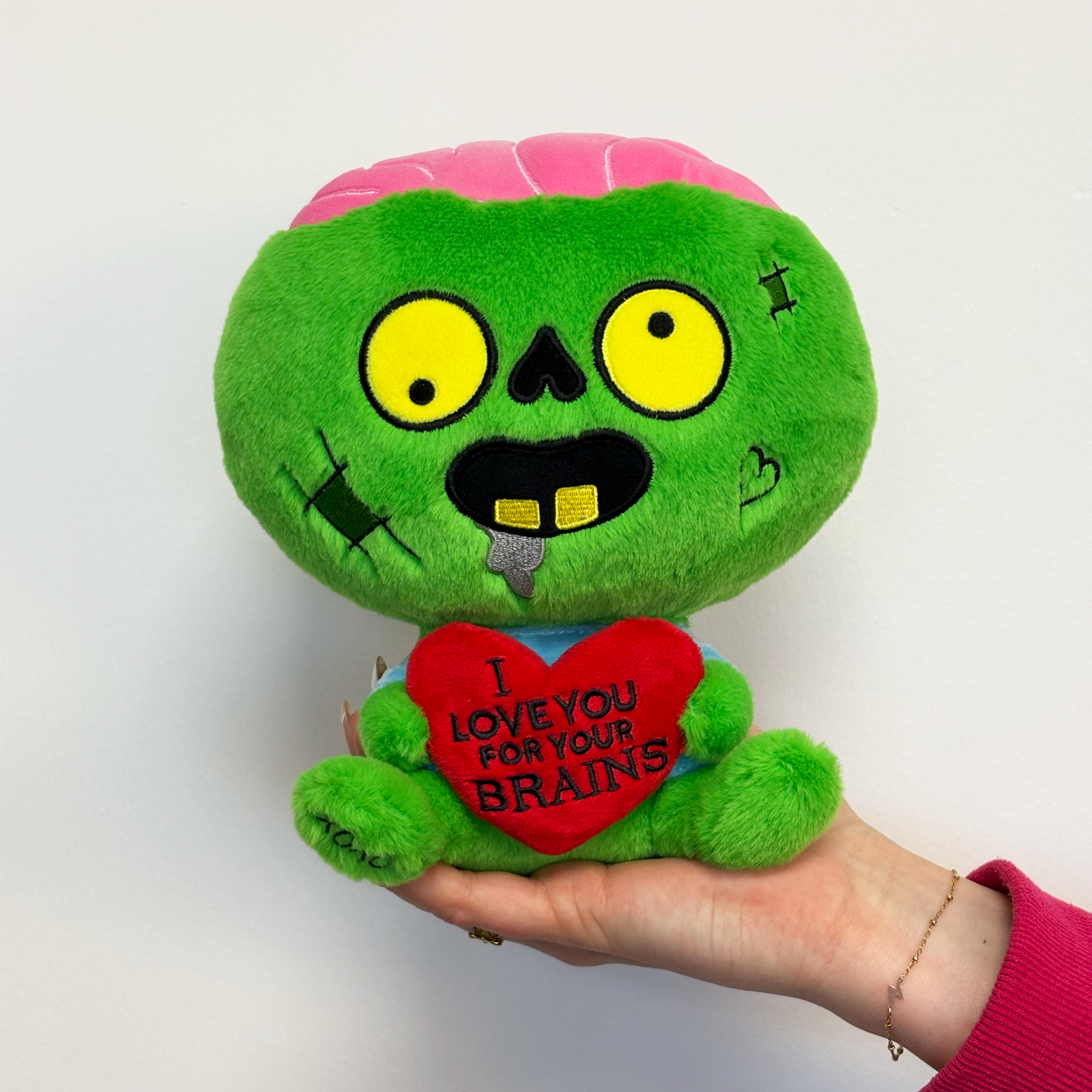 A hand holding a zombie plush with a 3D heart that reads, "I love you for your brains." 